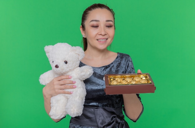 Happy asian woman holding teddy bear and box of chocolate candies as a gifts smiling cheerfully celebrating international women's day standing over green wall