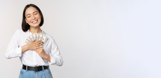 Счастливая азиатская женщина обнимает деньги в долларах и улыбается, стоя на белом фоне