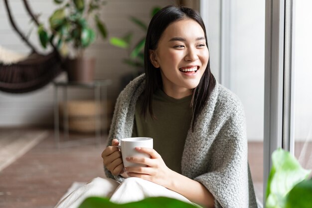 幸せなアジアの女性は、窓の近くの家の床に座って、週末の関係を楽しんで笑って笑っています...