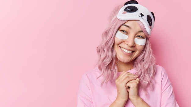 Happy Asian woman looks gladfully aside has dreamy expression keeps hands together smiles gently wears beauty patches wears sleepmask casual clothes isolated over pink background copy space.