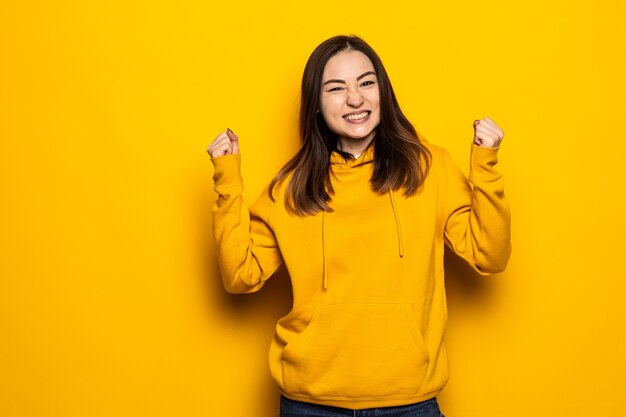 Happy asian woman make winning gesture isolated over yellow wall