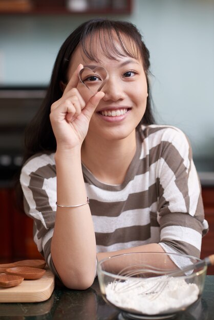 Happy Asian woman posing in kitchen and looking through hear-shaped cookie cutter