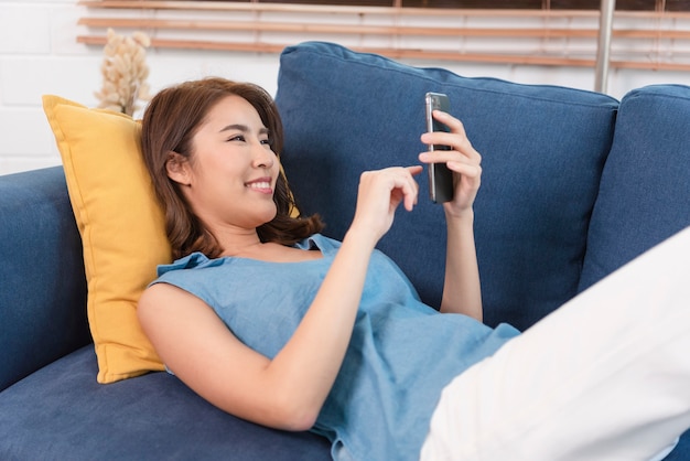 Felice donna asiatica che si distende sul comodo divano utilizzando smartphone in chat nei social network, guardando video divertenti a casa.