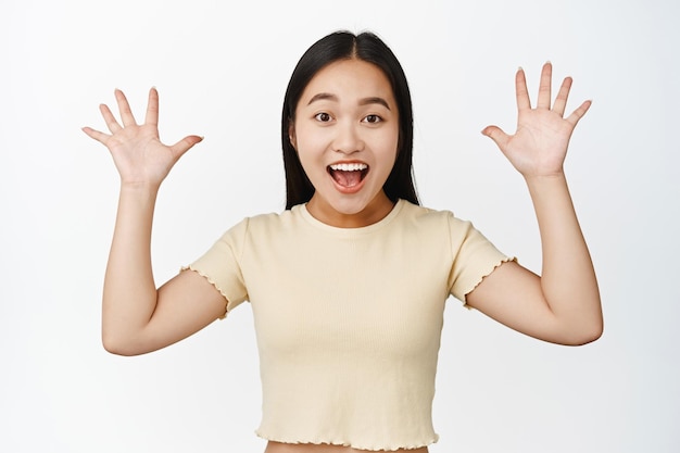 Foto gratuita donna asiatica felice che grida con le mani alzate dieci dita che sorridono eccitato in piedi in tshrit sopra priorità bassa bianca