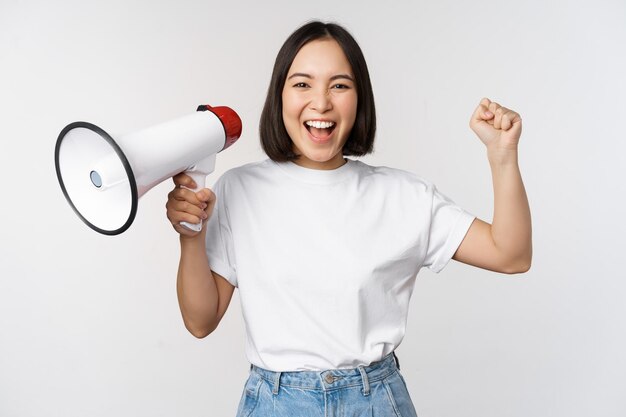 白い背景の上に立っている何かを宣伝するアナウンスを作るメガホンで叫んで幸せなアジアの女性