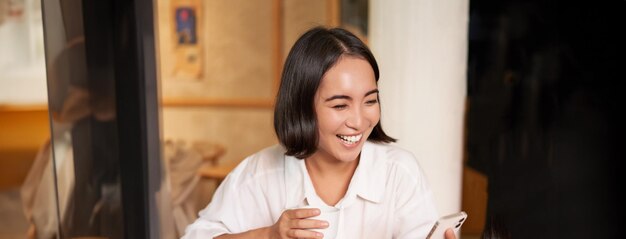 행복한 아시아 여성이 카페에 앉아 커피 한 잔을 마시고 스마트폰으로 비디오 통화에 대답하며 웃고 있습니다.