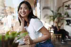 Free photo happy asian woman sitting in restaurant near window and smiling at camera.