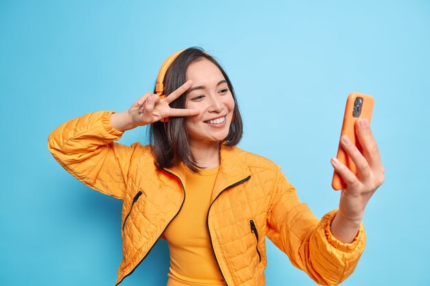 Happy Asian woman smiles gladfully makes peace gesture over eye takes selfie modern smartphone listens music via stereo wireless headphones isolated over blue wall. Lifestyle technology