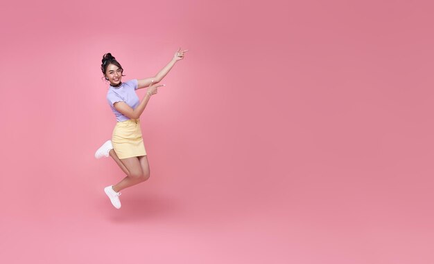 ピンクの背景のコピースペースに分離された手を指して笑顔とジャンプ幸せなアジアの女性