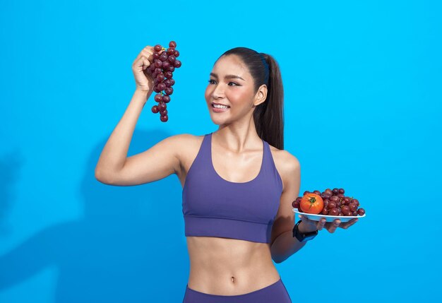 Счастливая азиатка в спортивной одежде ест свежие фрукты, набирает энергию для тренировки на синем фоне