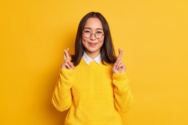 Happy Asian woman stands with crossed fingers believes in good luck on exam hopes dreams come true wears round spectacles and casual sweater.