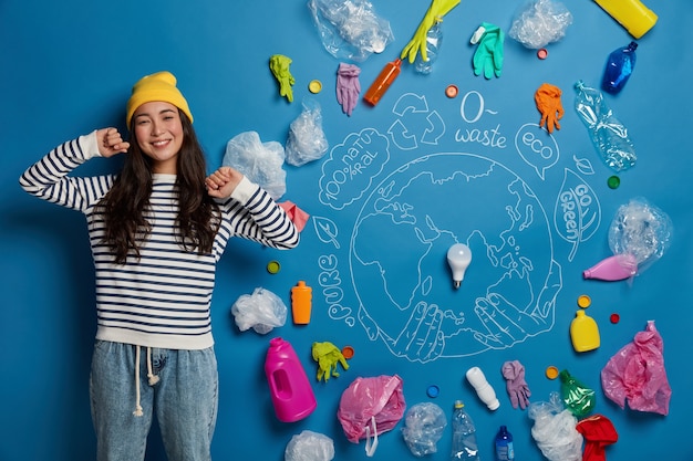 Free photo happy asian woman stretches hands, stands relaxed and satisfied, glad to finish cleaning territory from wastes, poses against blue background