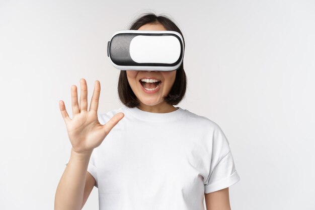 Happy asian woman using VR headset waving raised hands and laughing using virtual reality glasses standing over white background