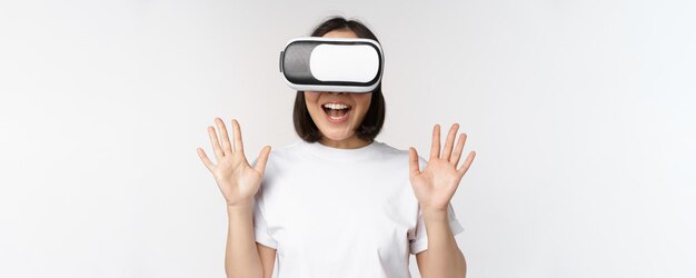 Happy asian woman using VR headset waving raised hands and laughing using virtual reality glasses standing over white background