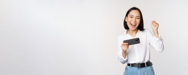 행복한 아시아 여성은 스마트폰 수평 위치를 잡고 흰색 배경을 축하하는 휴대 전화 비디오 게임에서 승리한 춤을 추며 승리합니다.