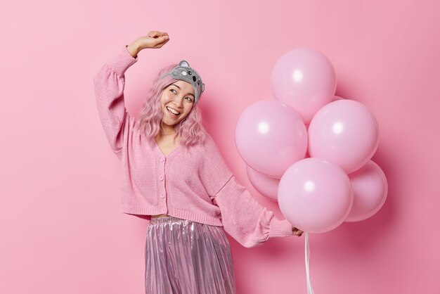 Счастливая азиатка с крашеными волосами в хорошем настроении носит повязку на лбу, джемпер и юбка беззаботно танцует, веселится на вечеринке, держит кучу надутых гелиевых шаров, изолированных на розовом фоне