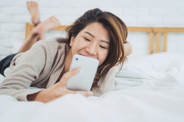 幸せなアジアの女性が午前中にベッドでスマートフォンを使用しています