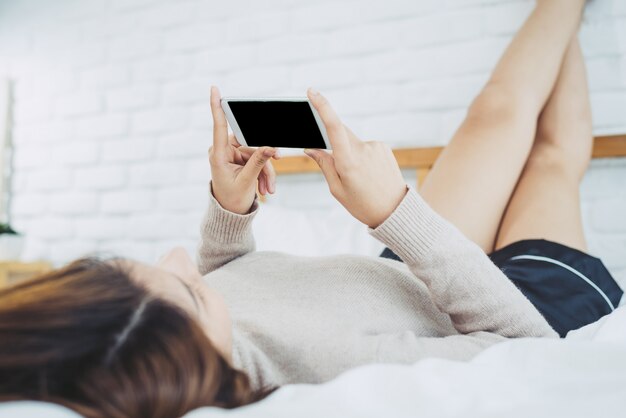 행복 한 아시아 여자는 아침에 침대에 검은 빈 빈 화면이 스마트 폰을 사용하고 있습니다