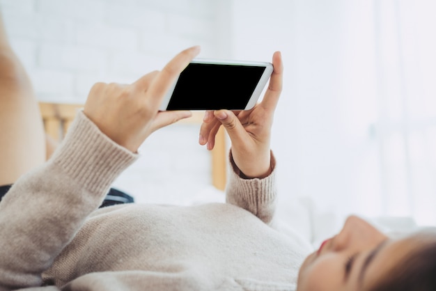 幸せな​アジア​の​女性​は​午前中​に​ベッド​に​黒い​空白​の​空​の​画面​を​持つ​スマート​フォン​を​使用しています