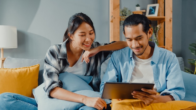 Happy asian young attractive couple man and woman sit on couch use tablet for shopping online furniture in new house