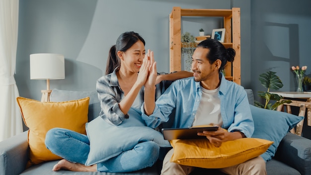 Happy asian young attractive couple man and woman sit on couch use tablet for shopping online furniture in new house