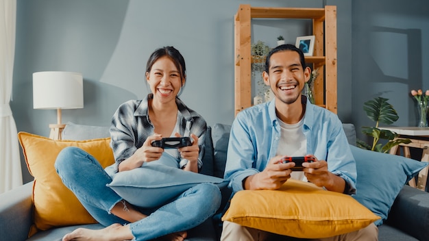 Happy asian young couple man and woman sit on couch use joystick controller for play video game