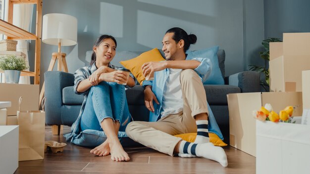 Happy asian young couple man and woman sit at new home drink coffee and talk with carton package box storage