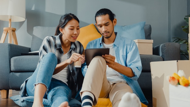 幸せなアジアの若いカップルの男性と女性は、新しい家のカートンパッケージで家を飾るためにオンラインショッピング家具にタブレットを使用しています。