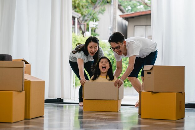 Happy Asian young family having fun laughing moving into new home. Japanese parents mother and father smiling helping excited little girl riding sitting in cardboard box. New property and relocation.