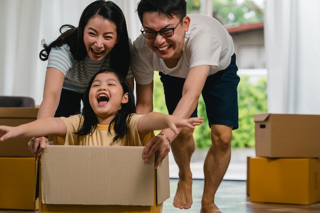 Happy Asian young family having fun laughing moving into new home. Japanese parents mother and father smiling helping excited little girl riding sitting in cardboard box. New property and relocation.