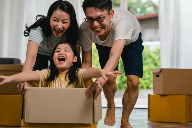 新しい家に移動して笑って楽しんで幸せなアジアの若い家族。日本人の親の母親と父親は、段ボール箱に座っている小さな女の子に乗って興奮して支援を笑っています。新しいプロパティと移転。