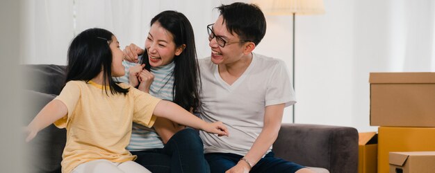 Happy Asian young family homeowners bought new house. Japanese Mom, Dad, and daughter embracing looking forward to future in new home after moving in relocation sitting on sofa with boxes together.