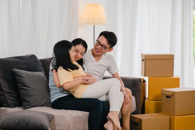 Happy Asian young family homeowners bought new house. Japanese Mom, Dad, and daughter embracing looking forward to future in new home after moving in relocation sitting on sofa with boxes together.