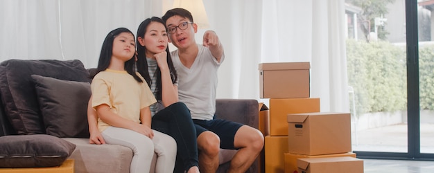 Happy Asian young family homeowners bought new house. Japanese Mom, Dad, and daughter embracing looking forward to future in new home after moving in relocation sitting on sofa with boxes together.