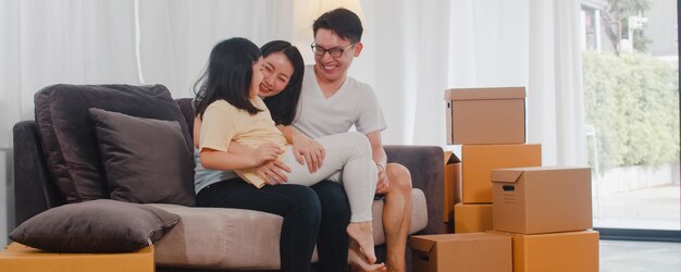 Happy asian young family homeowners bought new house. japanese mom, dad, and daughter embracing looking forward to future in new home after moving in relocation sitting on sofa with boxes together.