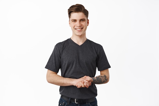 Happy to assist you. Smiling helpful young man holding hands together and looking friendly, listening to customer client, pleased to help, standing against white background
