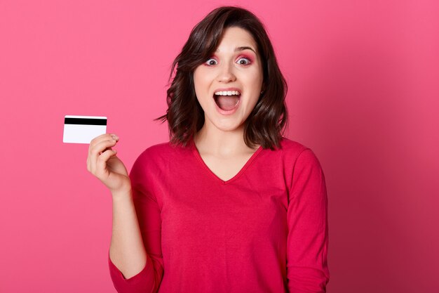 Happy astonished woman with credit card in hand, looking directly at camera with widely opened mouth and big eyes, brunette female win big sum of money.