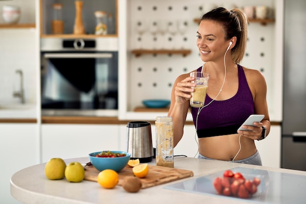 Счастливая спортивная женщина пьет смузи, используя мобильный телефон и слушая музыку через наушник на кухне