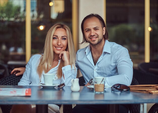 Счастливая привлекательная пара - очаровательная блондинка, одетая в белую блузку, и бородатый мужчина со стильной стрижкой, одетый в белую рубашку, во время свидания в кафе на открытом воздухе.