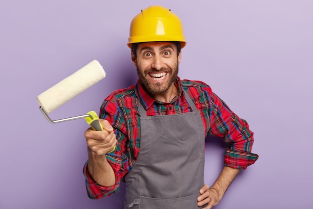 Felice e attraente caposquadra pronto per la riparazione della casa, tiene il rullo di vernice, decora le pareti, indossa un copricapo protettivo giallo, una camicia a scacchi e un grembiule, sorride positivamente. uomo con strumento di costruzione