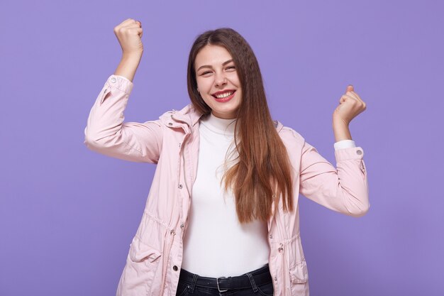 Happy attractive girl enjoys every moment of life, raises arms and clenches fists, has good mood, wears pale pink jacket and white turtleneck, isolated on lilac wall.