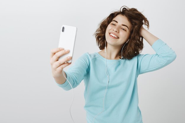 Happy attractive girl taking selfie on smartphone, posing with earphones