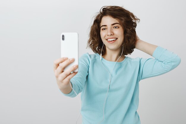 スマートフォンでselfieを取って、イヤホンでポーズをとって幸せな魅力的な女の子