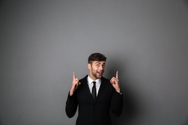 Happy attractive guy in black suit pointing with two fingers upward