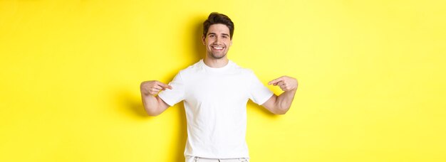 Happy attractive guy pointing fingers at your logo showing promo on his tshirt standing over yellow