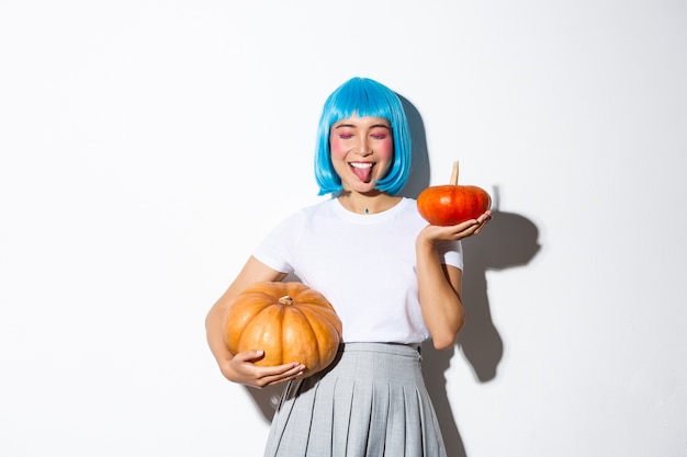 Happy attractive japanese girl in blue party wig, close eyes and showing tongue joyfully, celebrating halloween, holding two pumpkins.