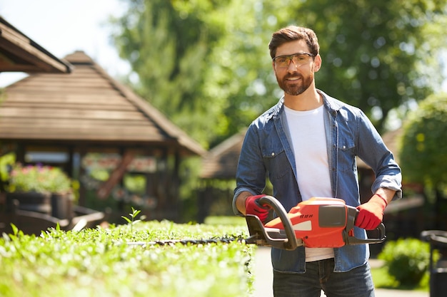 Foto gratuita giardiniere maschio attraente felice in vetri che posano mentre tagliano il cespuglio con le tagliasiepi elettriche