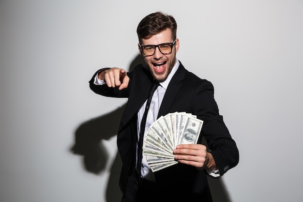 Happy attractive man in glasses and black suit holding bunch of money while pointing with finger on you