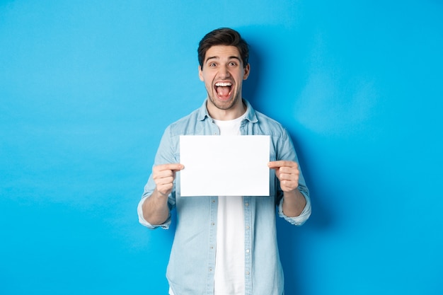 Happy attractive man showing piece of paper for your logo sign, standing amazed against blue background