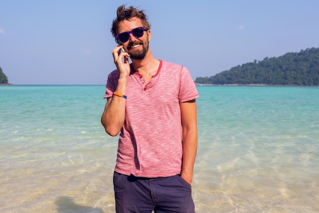 Happy attractive man in stylish sunglasses with beard using mobile phone at the beach